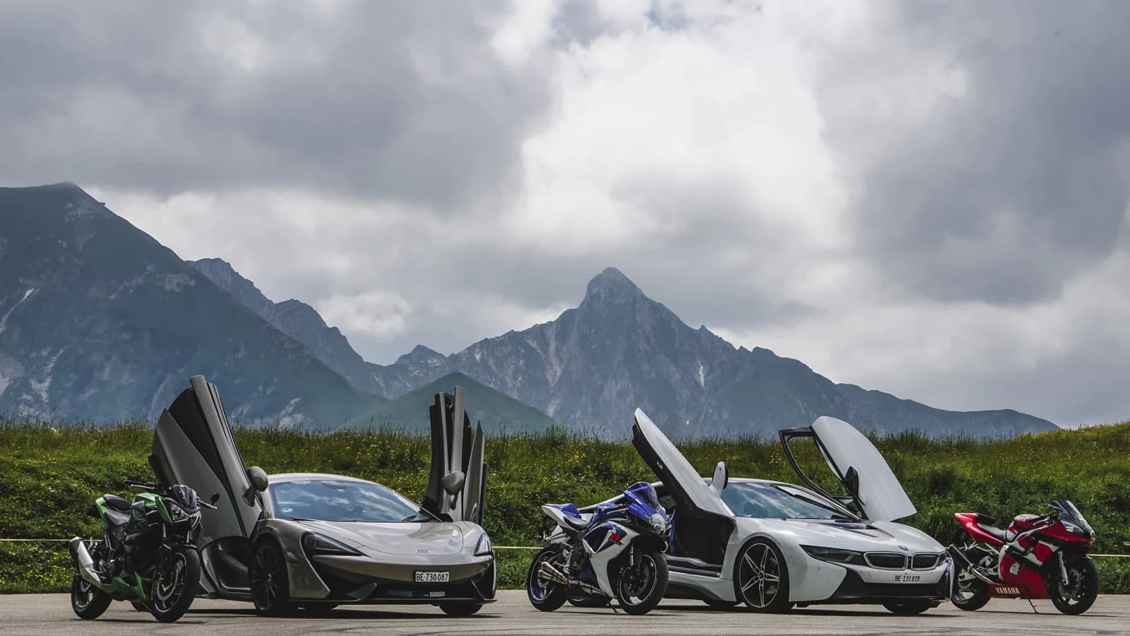 Sportwagen mit geöffneten Türen und Motorräder vor einer Berglandschaft